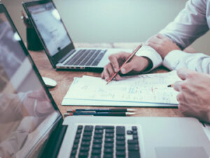 Closeup photo of a desk and two sets of hands. The desk has laptops and the hands are reviewing plans with a pencil