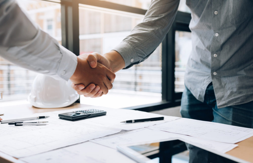 Two people shaking hands over a desk covered in construction plans