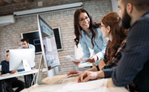 A woman stopping to talk to a man and woman sitting at a desk. She is showing the two of them papers