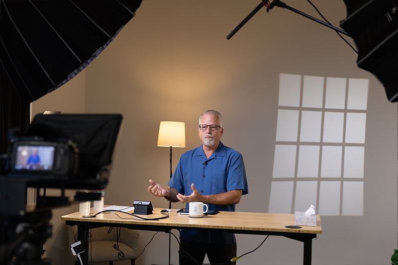 A man standing in front of a desk being filmed while teaching