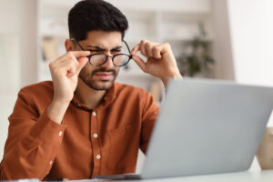 A man lowers his glasses and squints at his computer screen with a puzzled expression