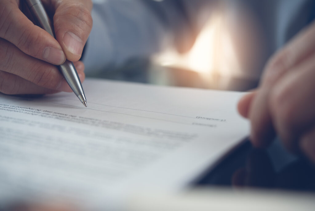 Closeup of a document with two hands, one is holding a pen to sign the document