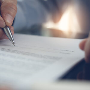 Closeup of a document with two hands, one is holding a pen to sign the document