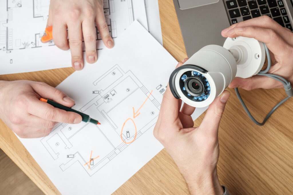 Top-down view of two sets of hands on a desk, one reviewing architecture plans and the other holding a security camera